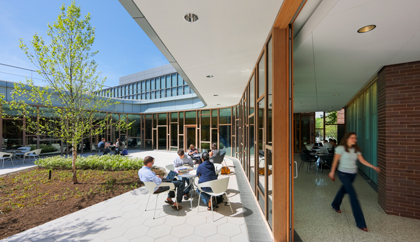 Students sit outside at tables next to a wall of windows, with a glass sliding door open, while others walk inside the building