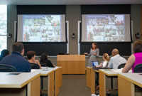 A professor lectures at the front of a full classroom that has side-by-side screens projecting images